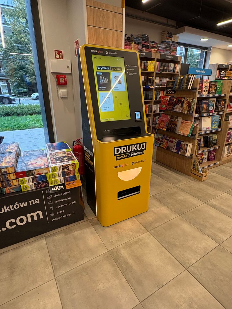 Żółto-czarny foto kiosk PhotoAiD do wydruku zdjęć i dokumentów A4 przy witrynie Empik na Woli